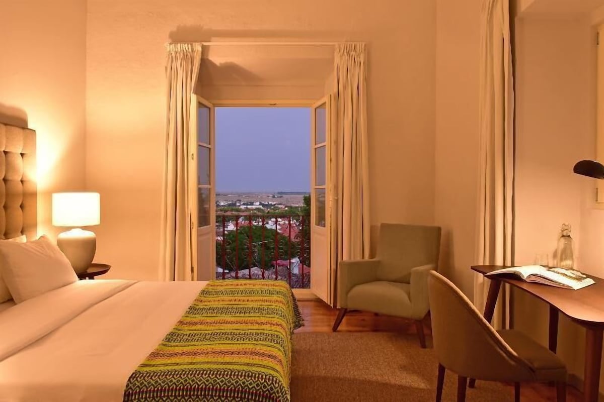 A hotel room with a bed, a desk, a chair, and a view of the cityscape.