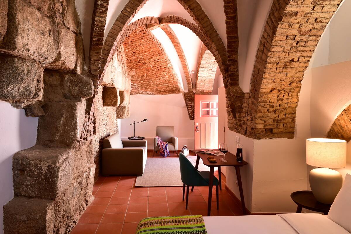 A cozy room with a stone wall, a bed, a desk with a lamp, and a chair.