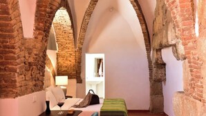 A room with exposed brick walls, a bed with pillows, a bedside table with a glass, and a lamp. - Noble House Essence Évora - by Unlock Hotels (Évora)