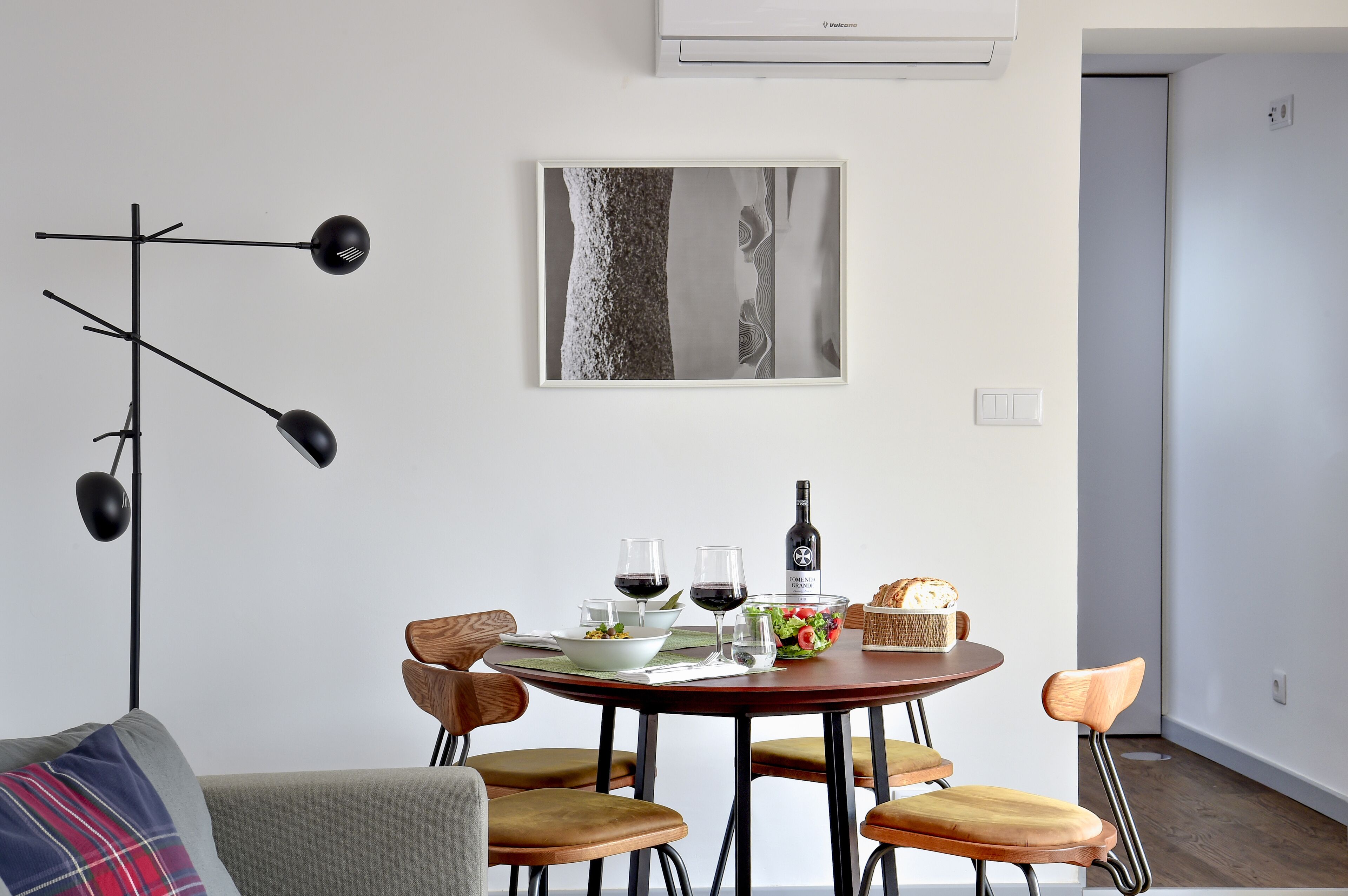 A modern dining area with a round table, four wooden chairs, a black floor lamp, and a framed abstract artwork on the wall.