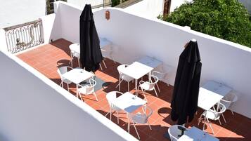 A rooftop terrace with white tables and chairs, black umbrellas, and a red-tiled floor.