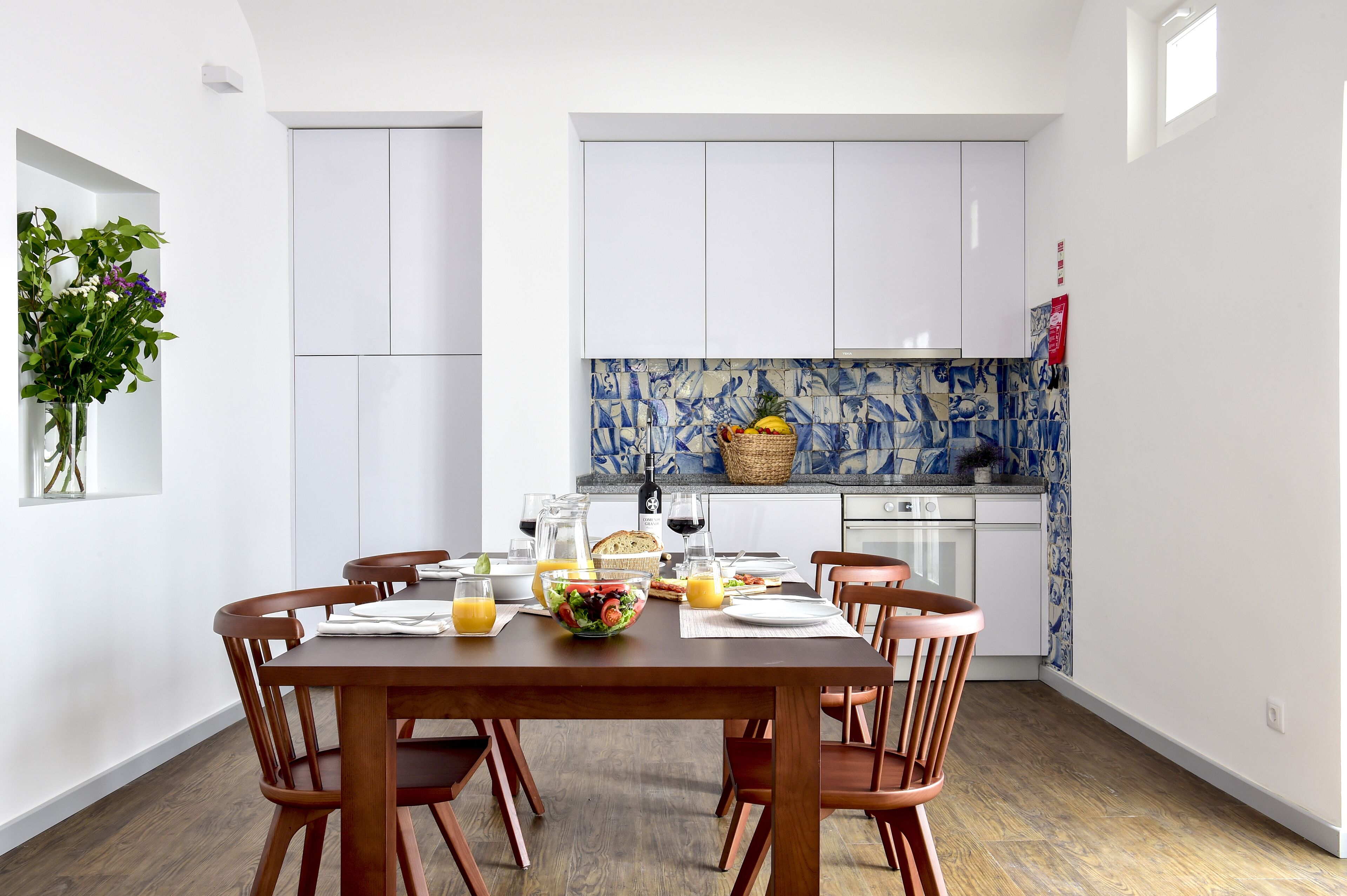 A dining area with a wooden table set for four, surrounded by chairs. A kitchen with white cabinets and blue patterned backsplash is visible in the background.