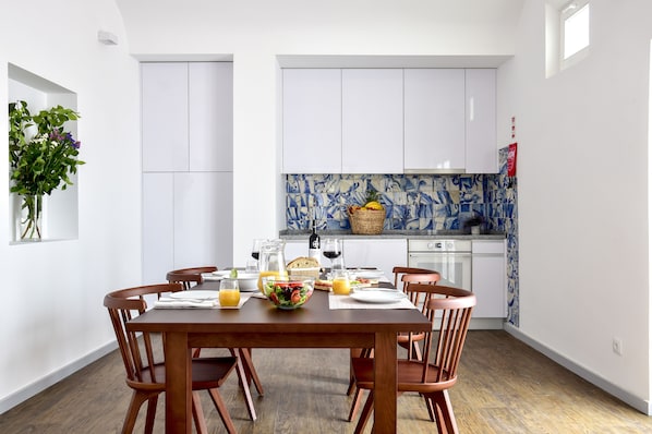 A dining area with a wooden table set for four, surrounded by chairs. A kitchen with white cabinets and blue patterned backsplash is visible in the background. - Noble House Essence Évora - by Unlock Hotels (Évora)