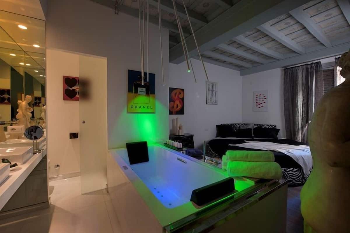 A modern bathroom with a glass-enclosed shower, a large mirror, and a bed with black and white bedding in the adjacent room.