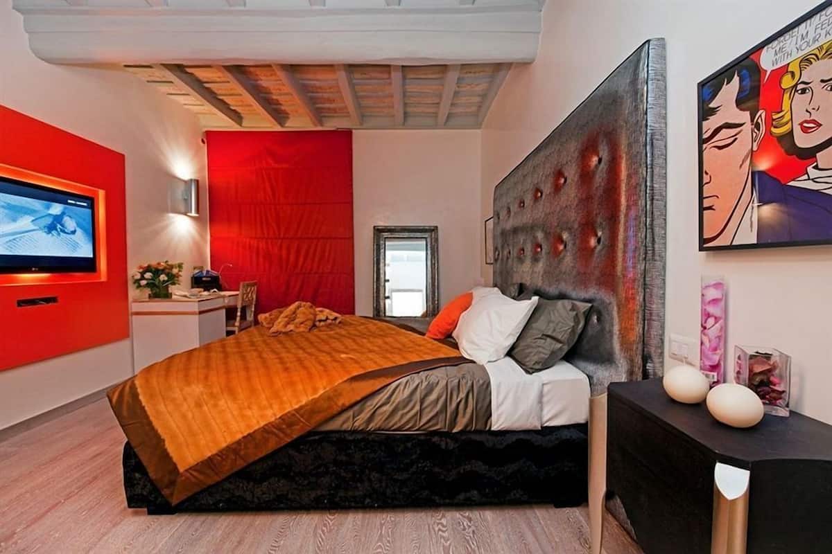 A modern bedroom with a red accent wall, a large bed with a unique headboard, a flat-screen TV, and a painting on the wall.