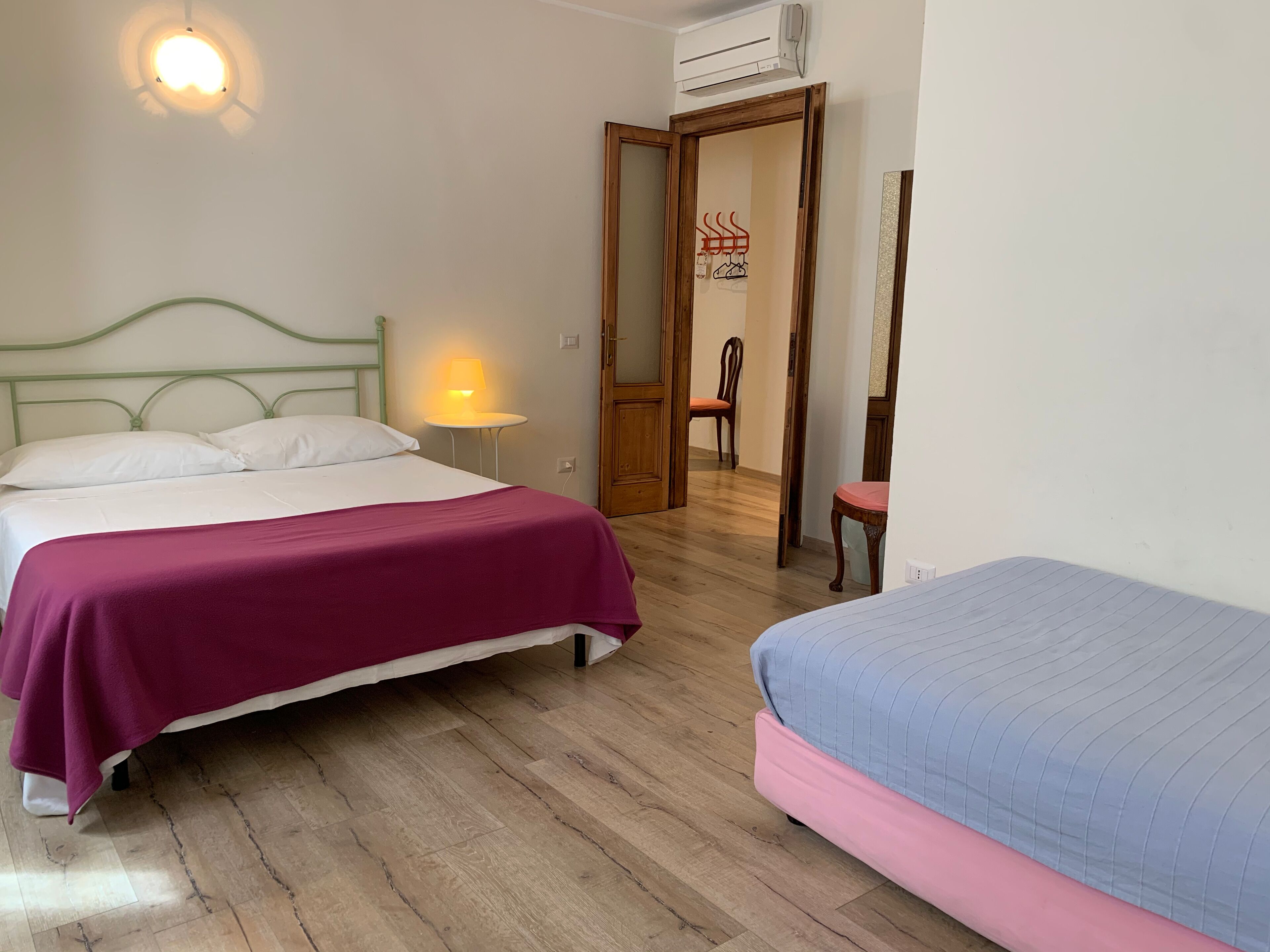Una habitación con una cama, una mesita de noche con lámpara, una silla y una puerta abierta que da a otra habitación.