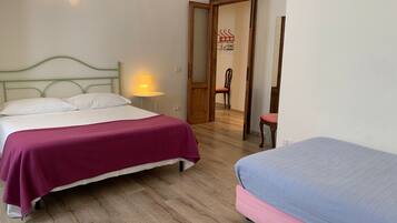 Una habitación con una cama, una mesita de noche con lámpara, una silla y una puerta abierta que da a otra habitación.