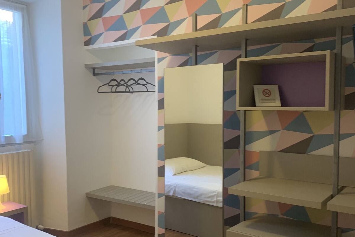 Une chambre de dortoir équipée d’un lit superposé, d’un bureau, d’une chaise, d’une fenêtre avec des rideaux et d’un mur orné de motifs géométriques.