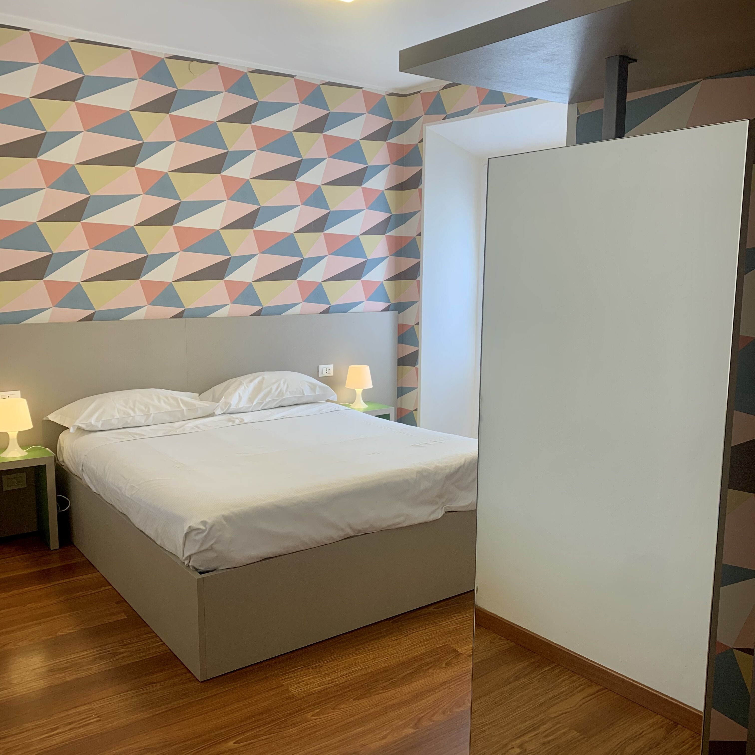 Un dormitorio con una cama, dos mesitas de noche con lámparas, una pared con estampado geométrico y suelo de madera.