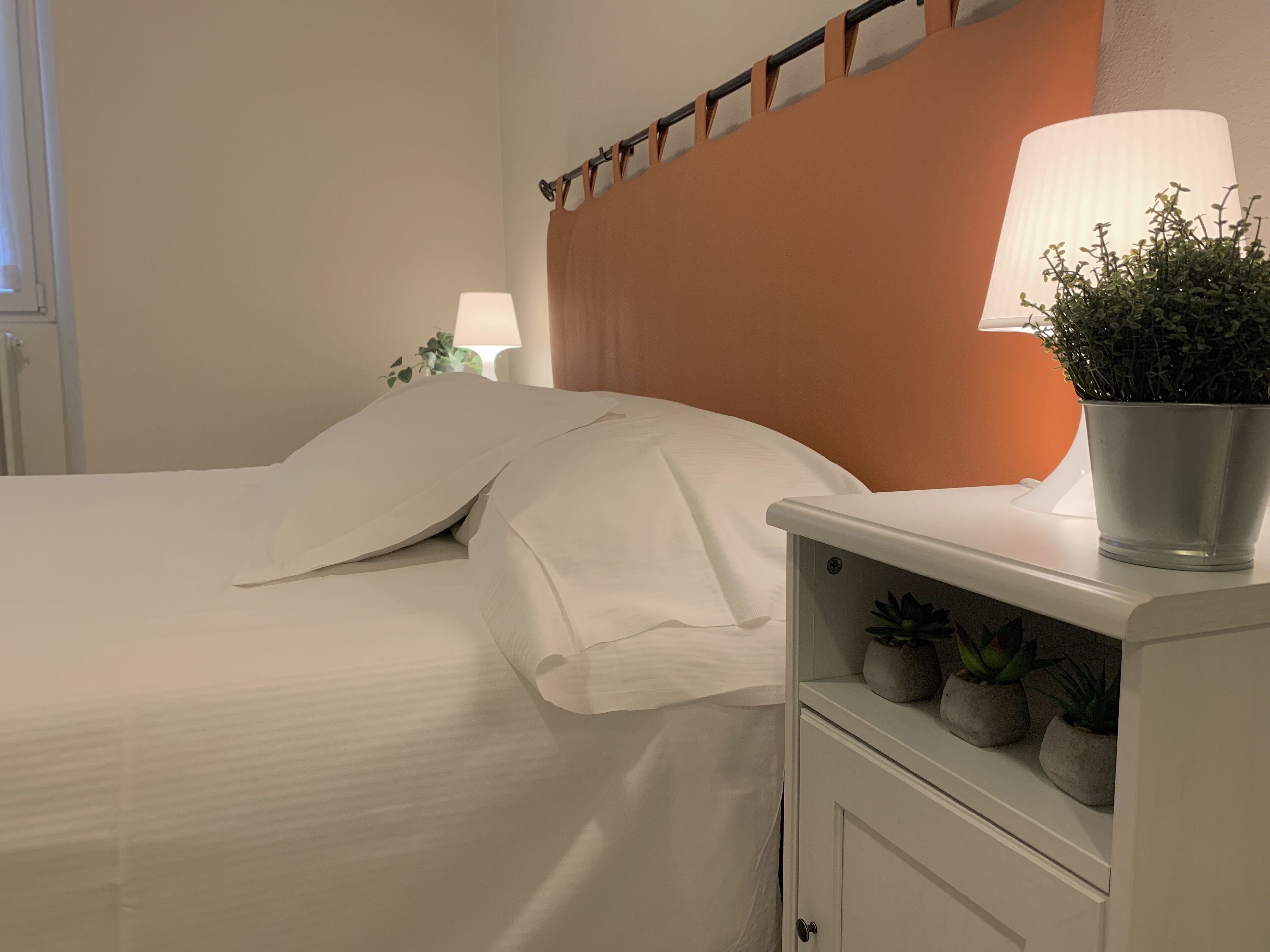 A neatly made bed with white linens, a bedside lamp, and a shelf with potted plants.