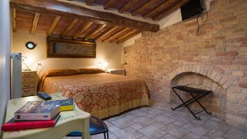 A bedroom with a brick fireplace, a bed with a floral bedspread, a desk with books and a remote control, and a wooden ceiling.