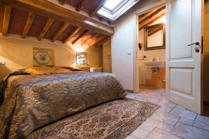 A bedroom with a bed, a bedside lamp, a rug, a skylight, and a bathroom with a sink and mirror. - Donna Nobile (San Gimignano)
