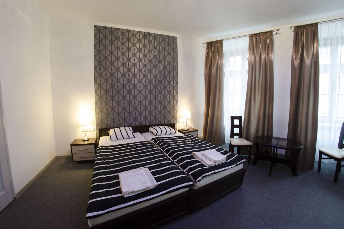 A double bed with a striped bedspread, two bedside tables with lamps, a small table with a chair, and a patterned wall.