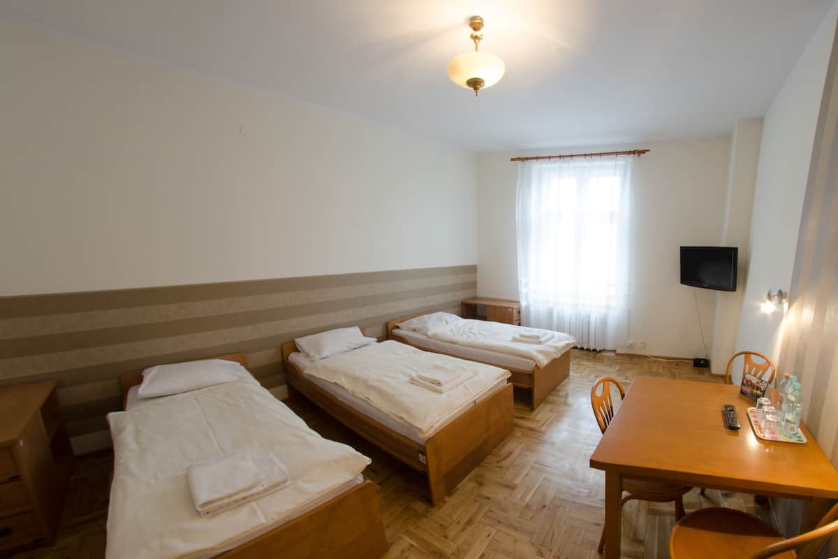 A hotel room with three single beds, a wooden table with chairs, a TV, and a window with curtains.
