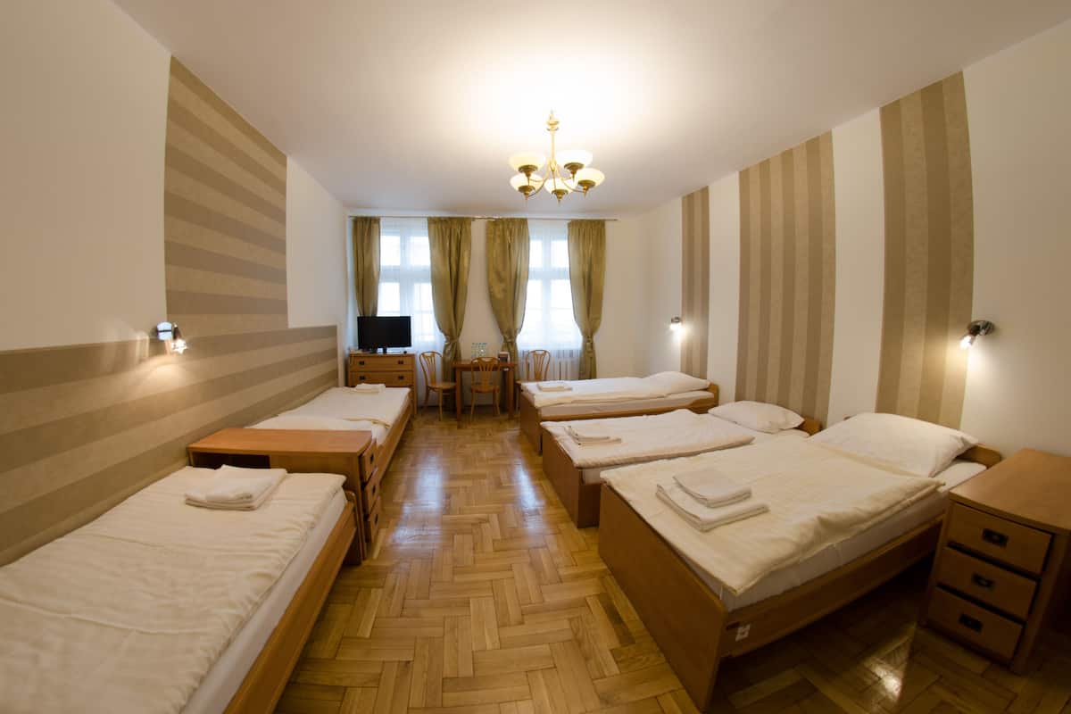 A hotel room with four single beds, a wooden floor, a desk with a chair, a television, and a chandelier.