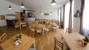 Zona pranzo con tavoli e sedie in legno, angolo cottura con frigorifero e scaffali, e ampie finestre con tende.