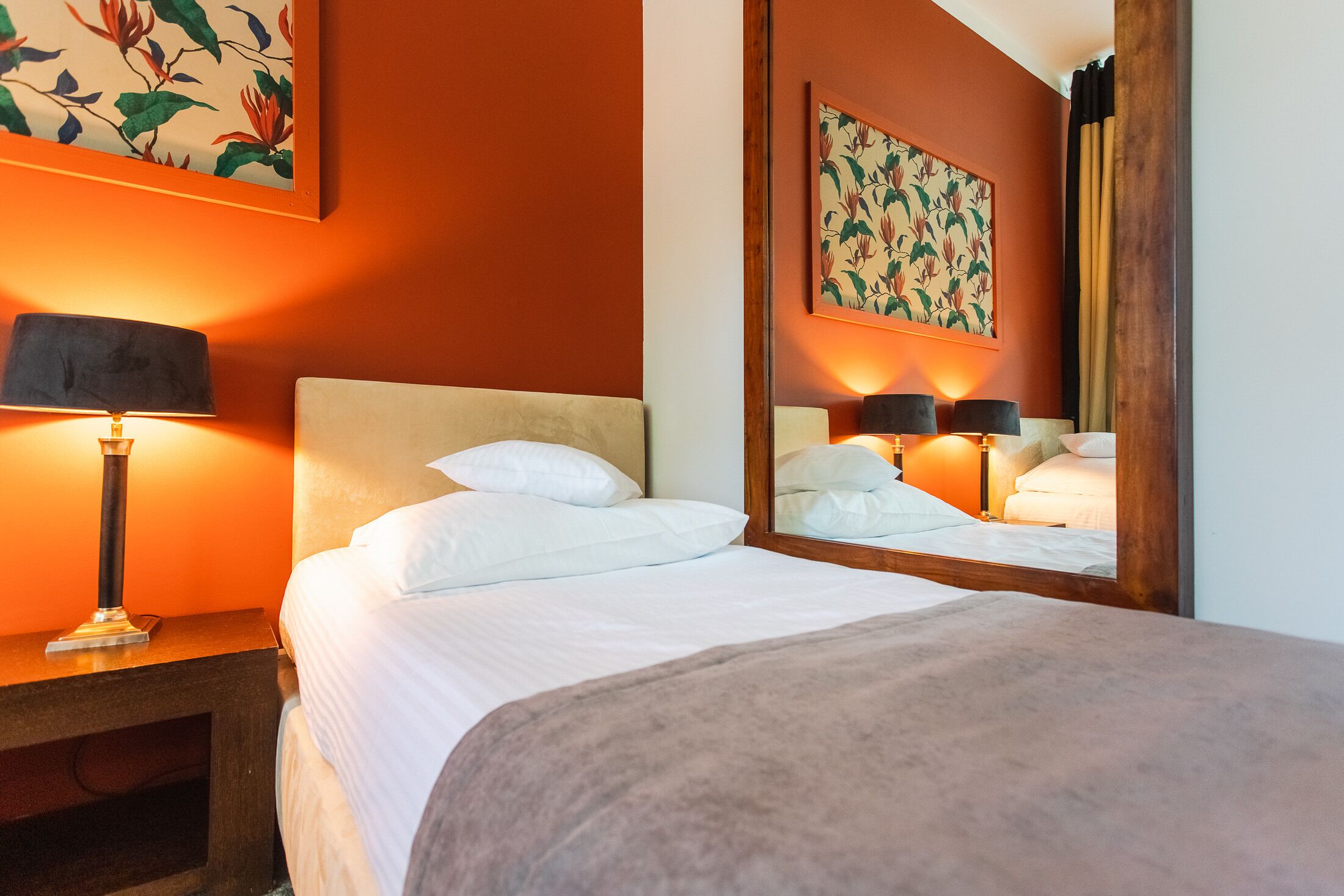 A hotel room with a bed, a nightstand with a lamp, and a large mirror reflecting the room.