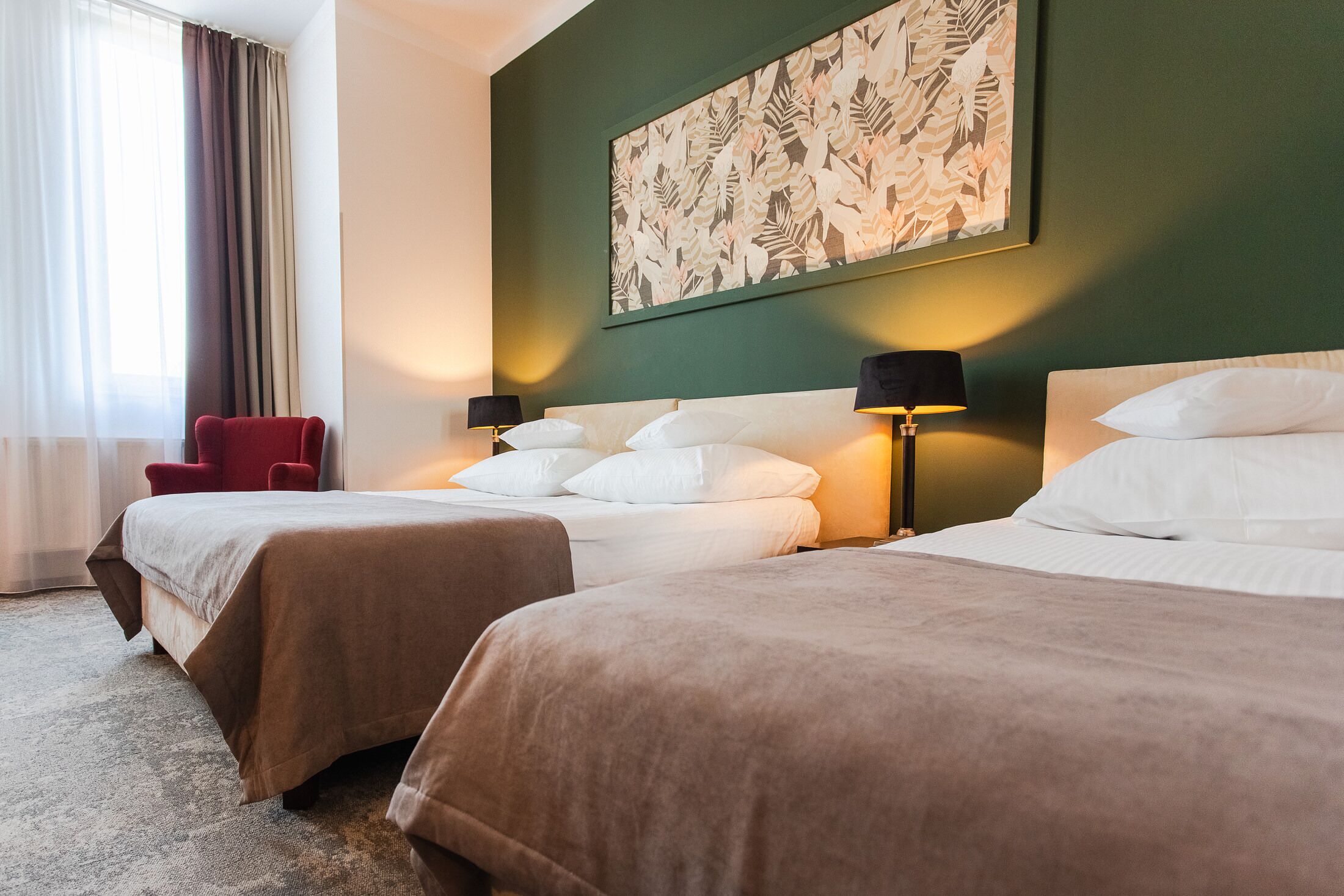 Habitación de hotel con dos camas, una silla roja, un cuadro floral grande en la pared y una ventana con cortinas.
