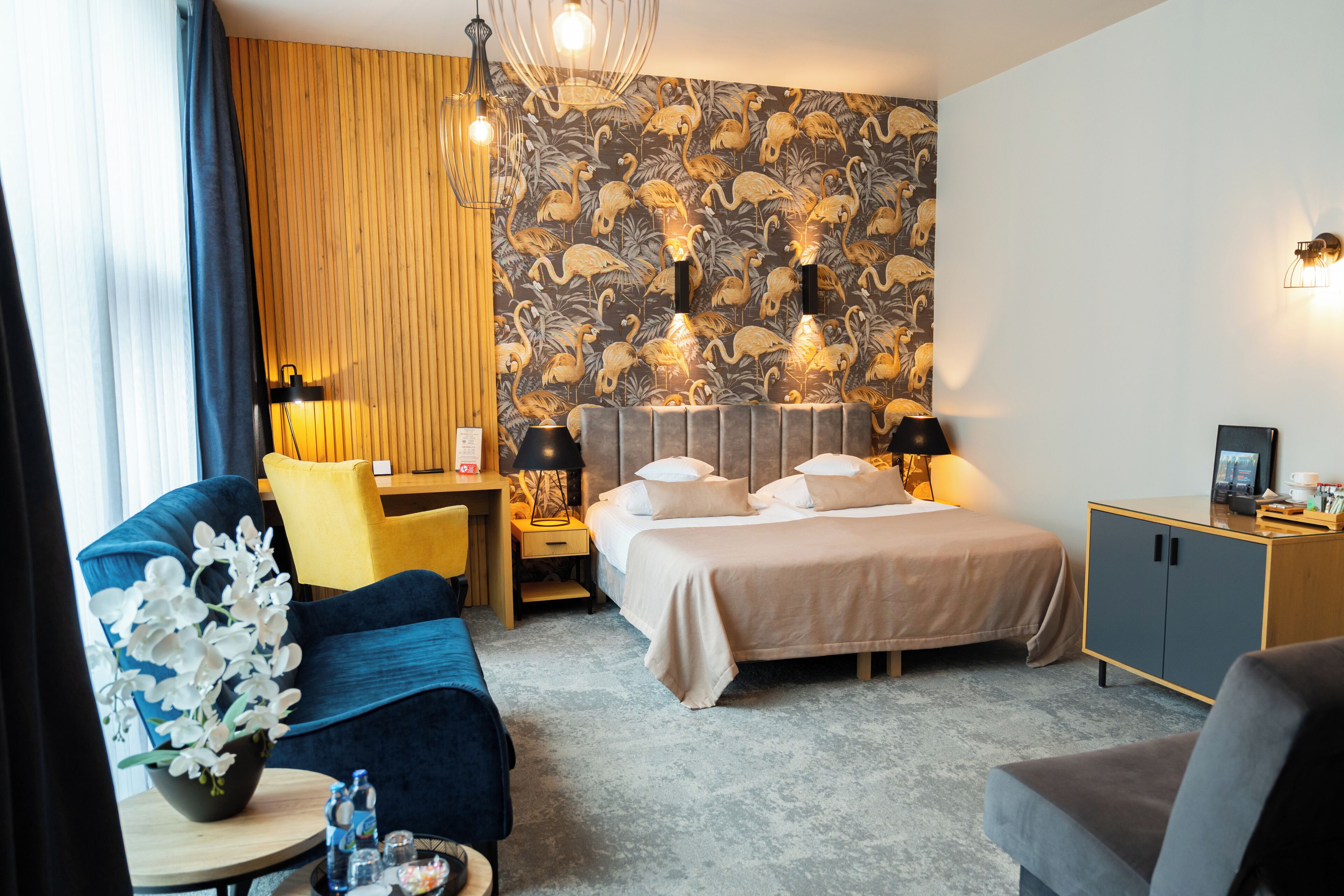 Ein Hotelzimmer mit Bett, Schreibtisch, Stuhl, Sofa, Lampe, einer Blumenvase und einer Wand mit Blumenmuster.