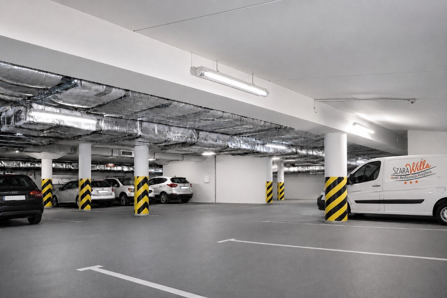 Un parking souterrain où sont garées plusieurs voitures, des piliers ornés de rayures jaunes et noires, et un fourgon blanc portant l’inscription « SZARA Willa ».
