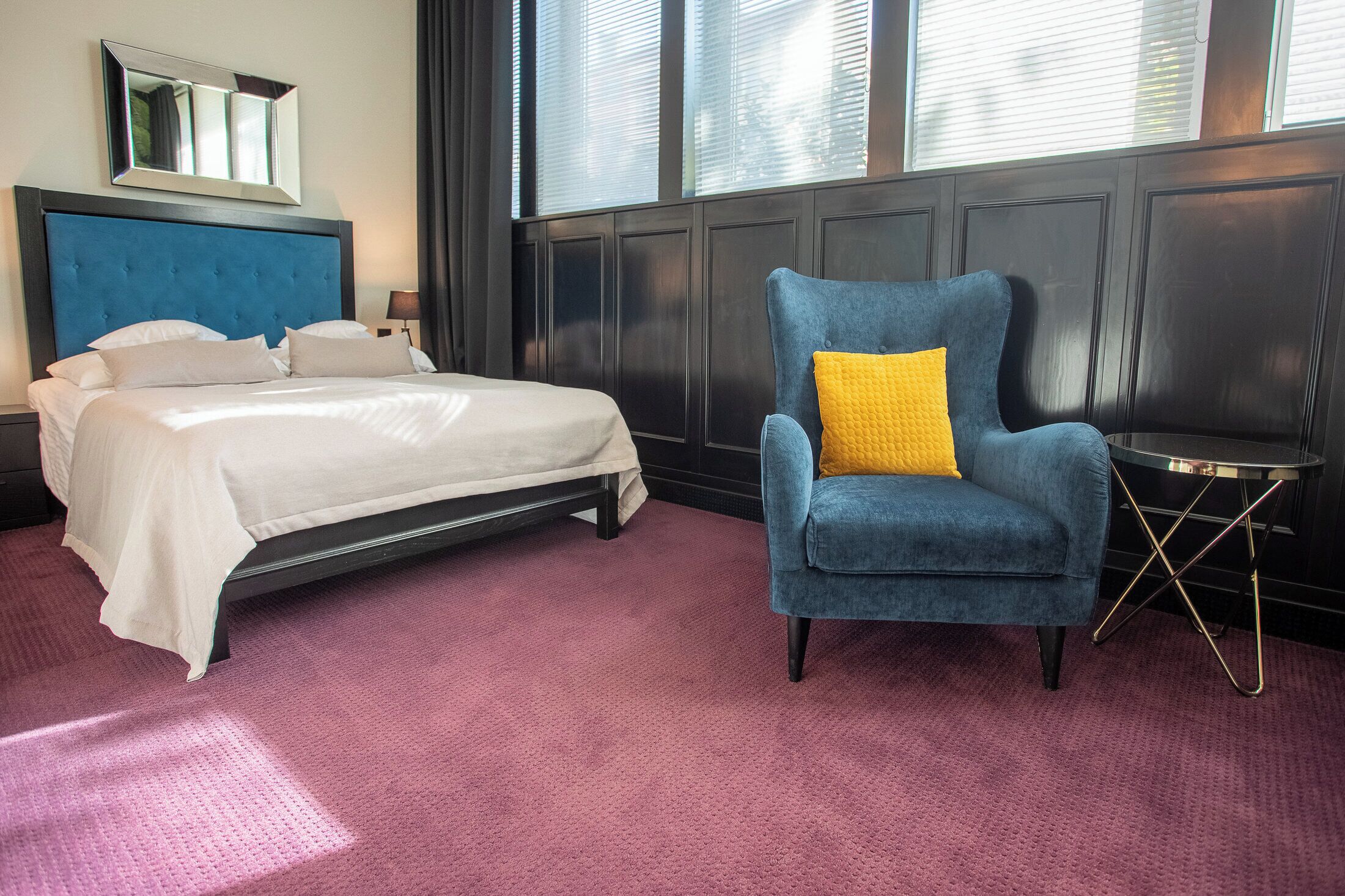A hotel room with a bed, a blue armchair with a yellow pillow, a small table, and a window with blinds.