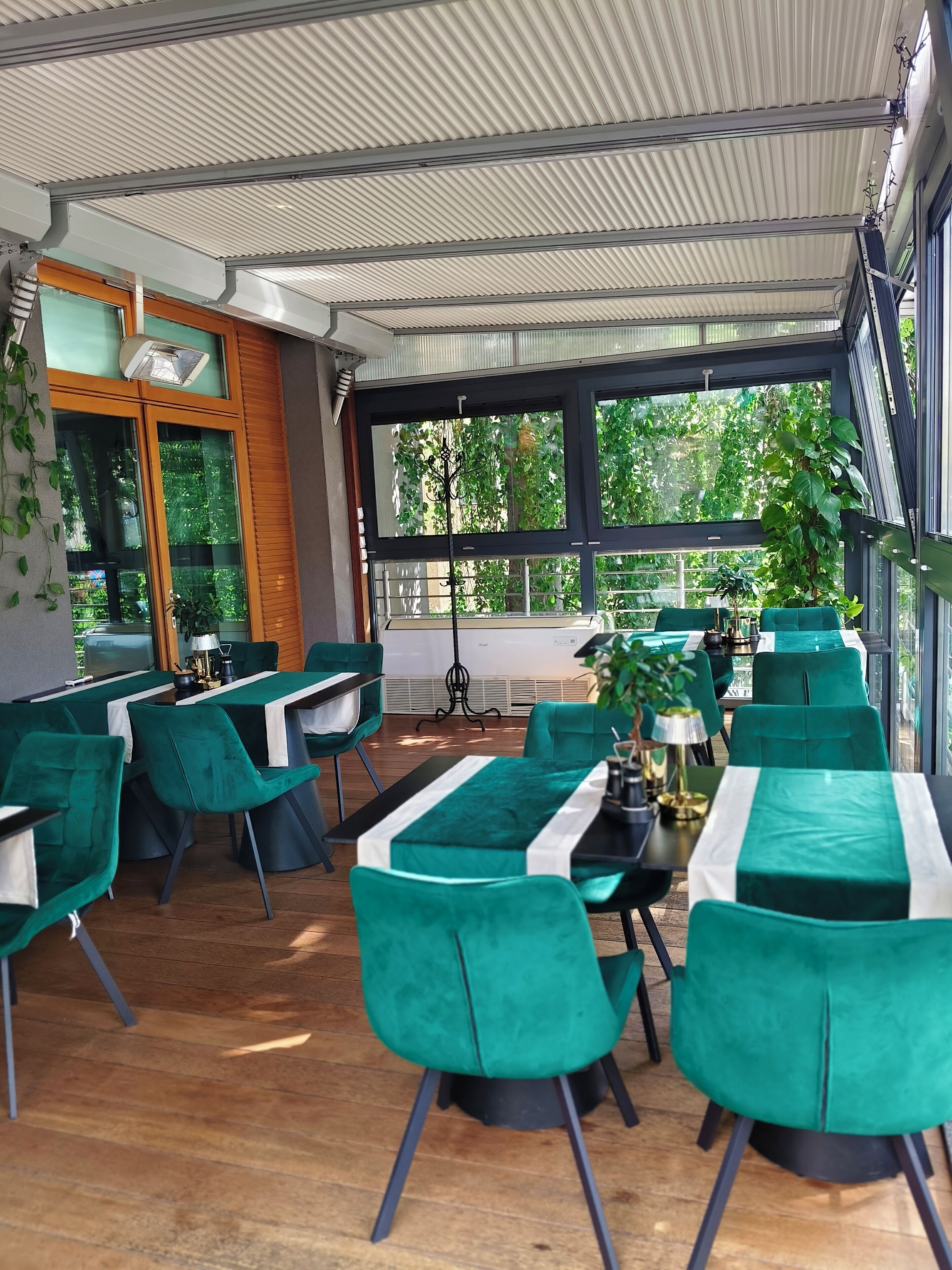A modern indoor dining area with green upholstered chairs, tables with white tablecloths, and large windows offering a view of greenery outside.