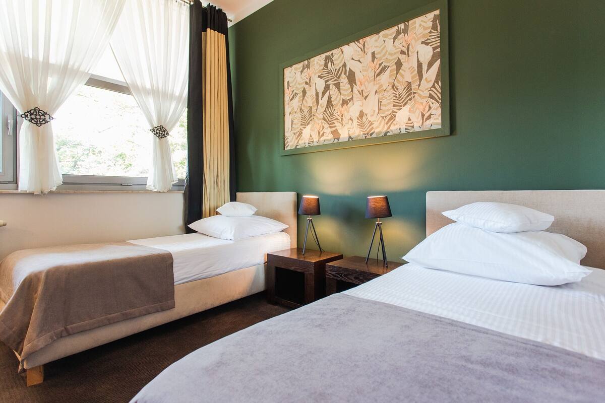 Habitación de hotel con dos camas, un ventanal con cortinas traslúcidas, una obra de arte en la pared y lámparas de noche.