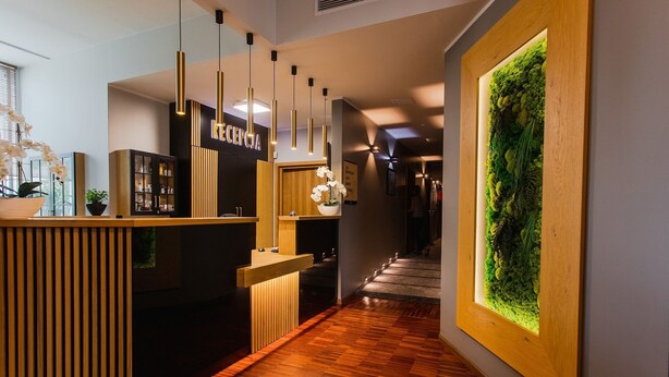 Un área de recepción con un mostrador de madera, iluminación colgante, una planta verde en la pared y un letrero con texto cirílico.