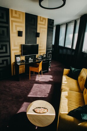 A modern hotel room with a dark patterned wall, a flat-screen TV, a desk with a chair, a sofa, and a small round table with a spoonful of food. - Szara Willa (Opole)