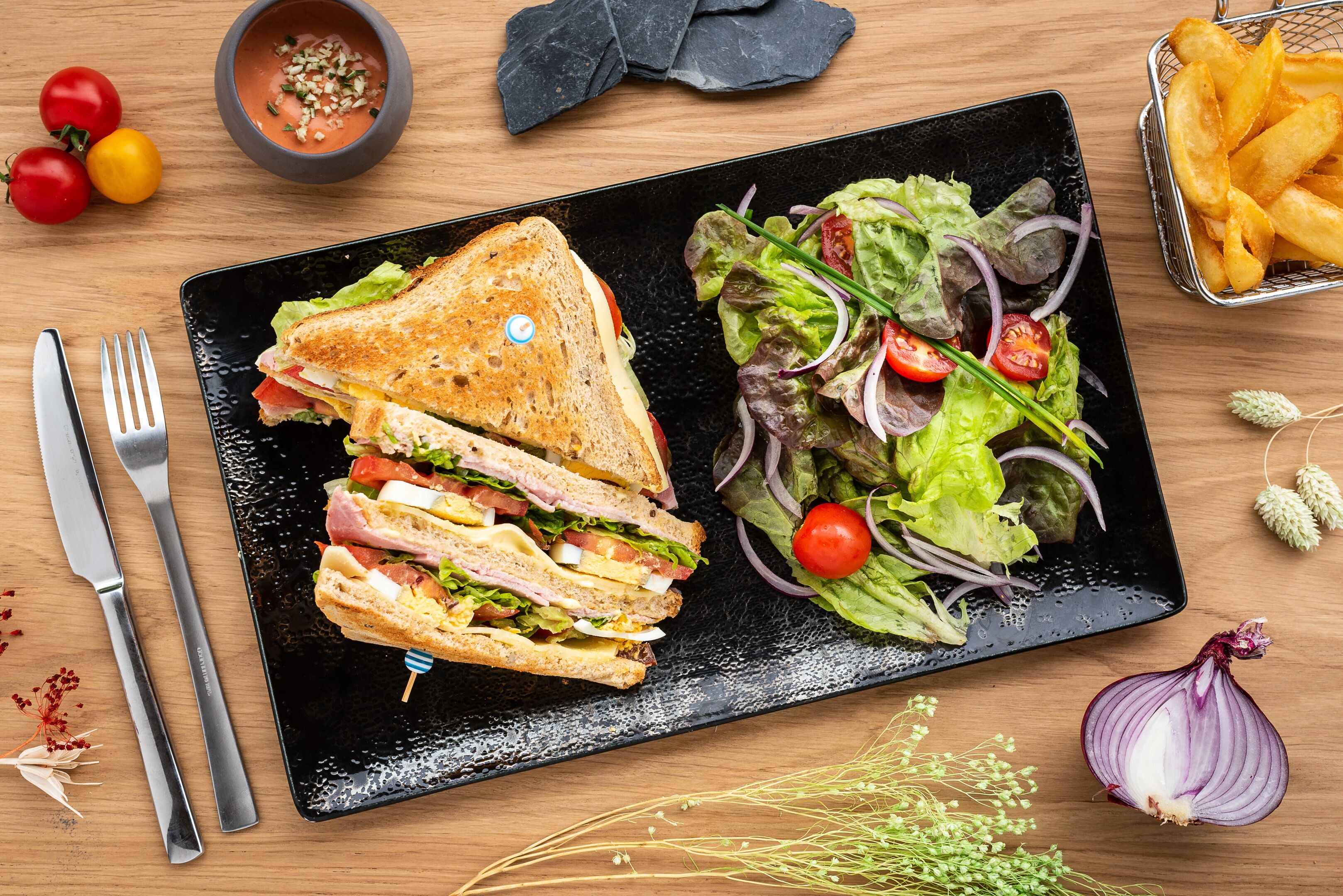 A black plate with a sandwich and salad, a bowl of sauce, and a basket of fries on a wooden table.