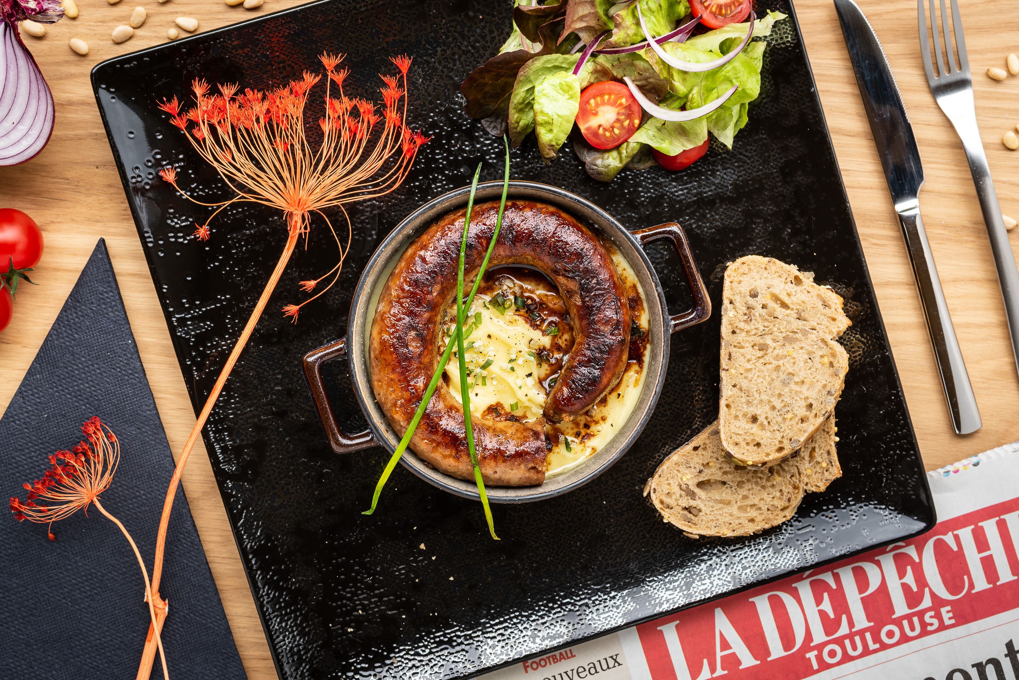 Un repas composé de saucisses, de pain et d’une salade, le tout servi dans une assiette noire.