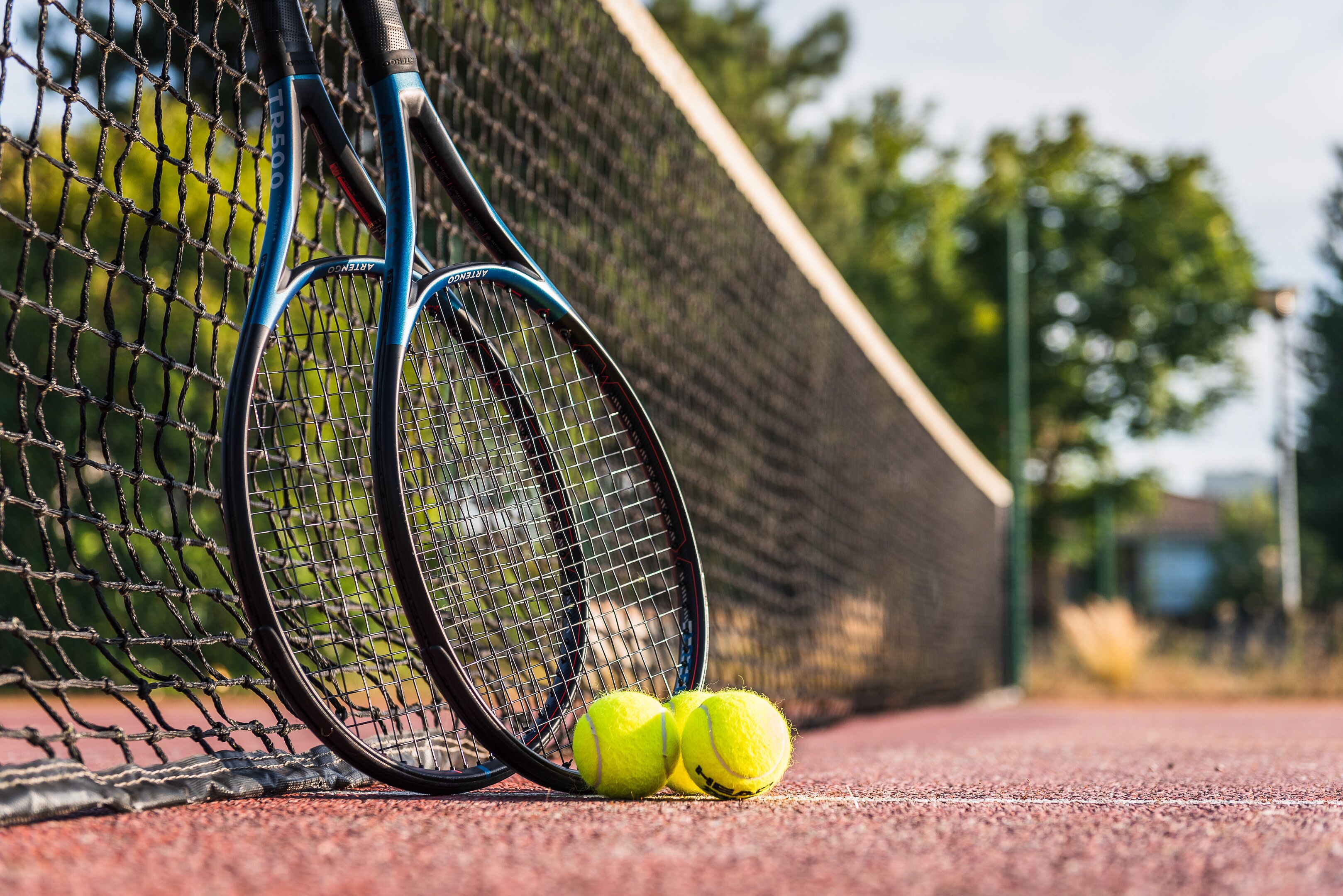 A tennis court with two tennis rackets leaning against the net and three tennis balls on the ground.