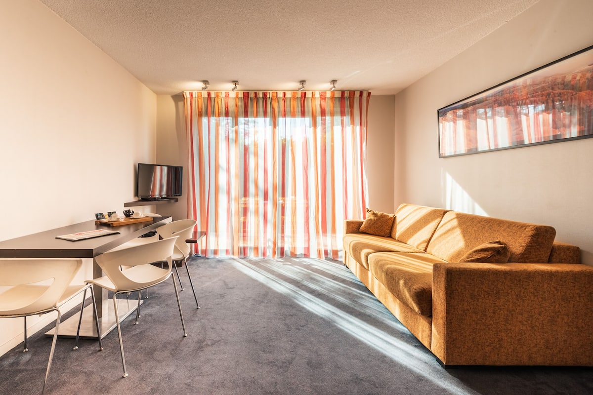 Une chambre d’hôtel moderne avec un tapis gris, un canapé, une télévision, un bureau avec des chaises et une grande fenêtre agrémentée de rideaux à rayures.