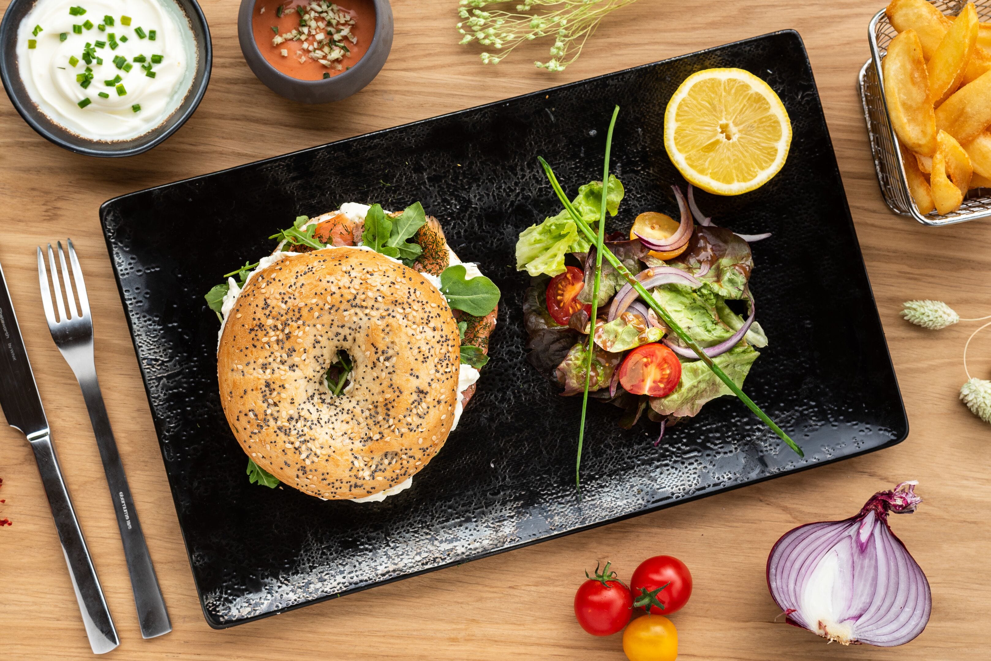 A black plate with a bagel sandwich and a salad on a wooden table.
