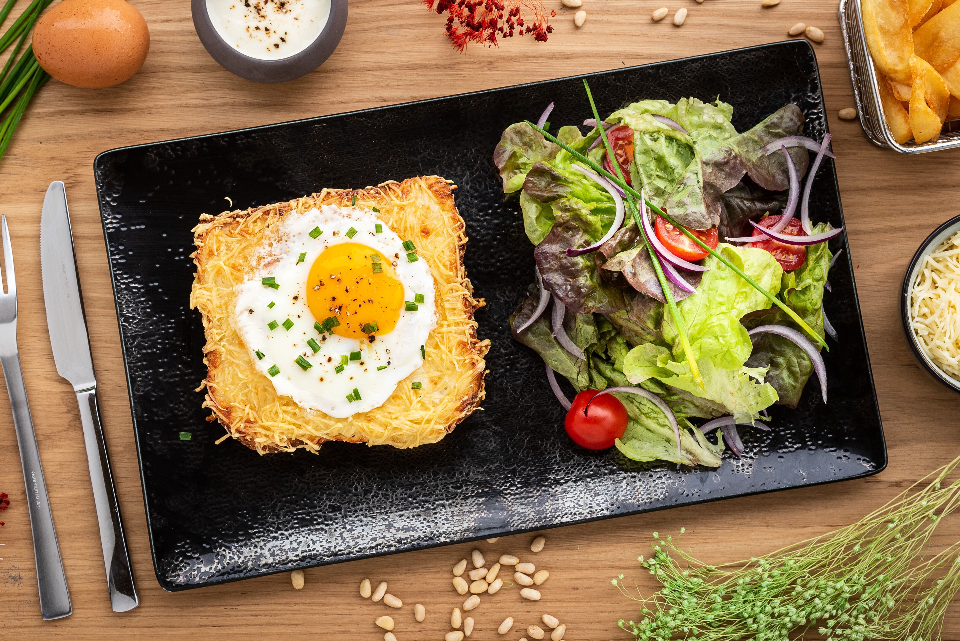 A black plate with a fried egg on a crispy base, a side salad, and a bowl of soup.