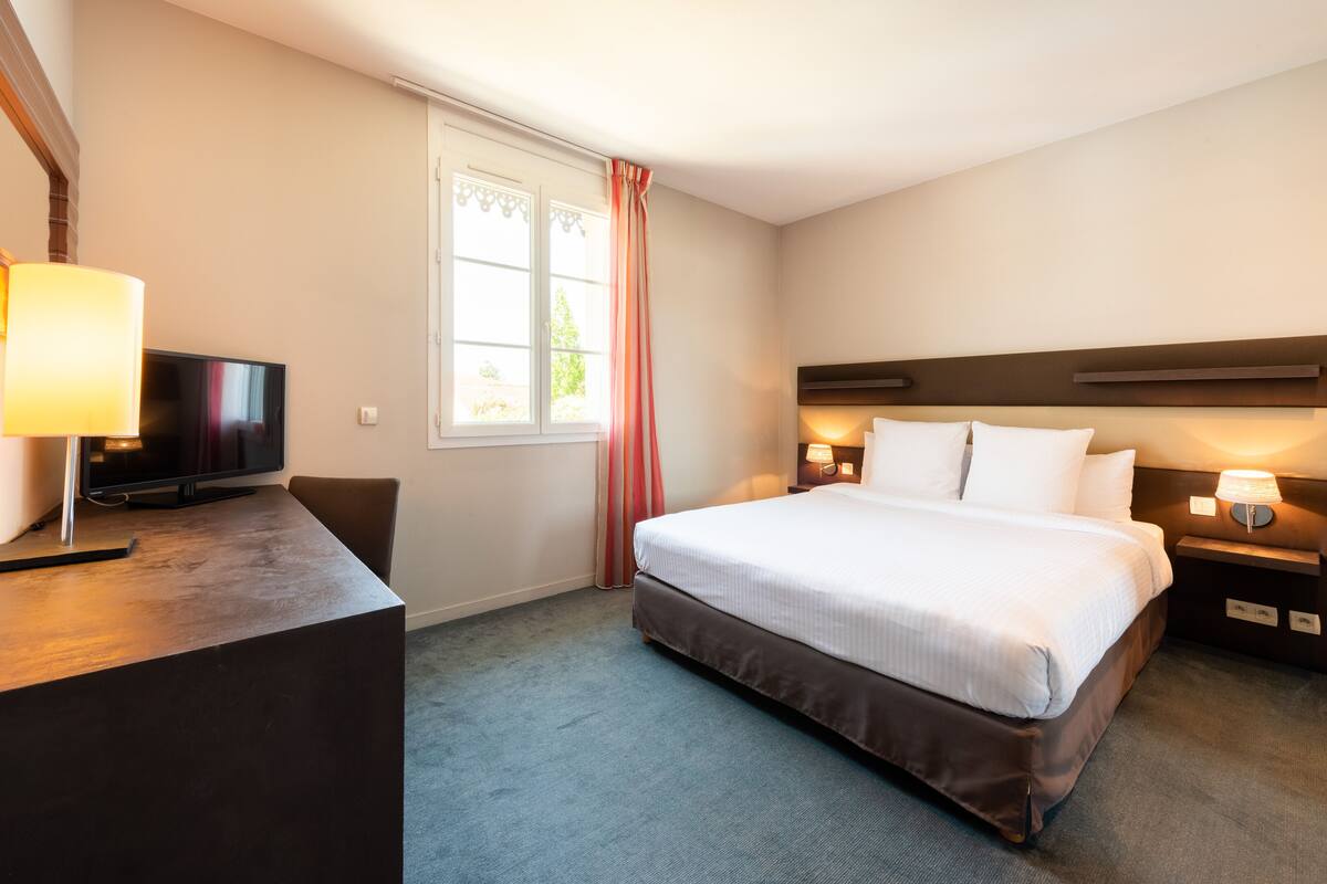Une chambre d’hôtel comprenant un lit, des lampes de chevet, un bureau avec une lampe, une télévision et une fenêtre avec des rideaux.