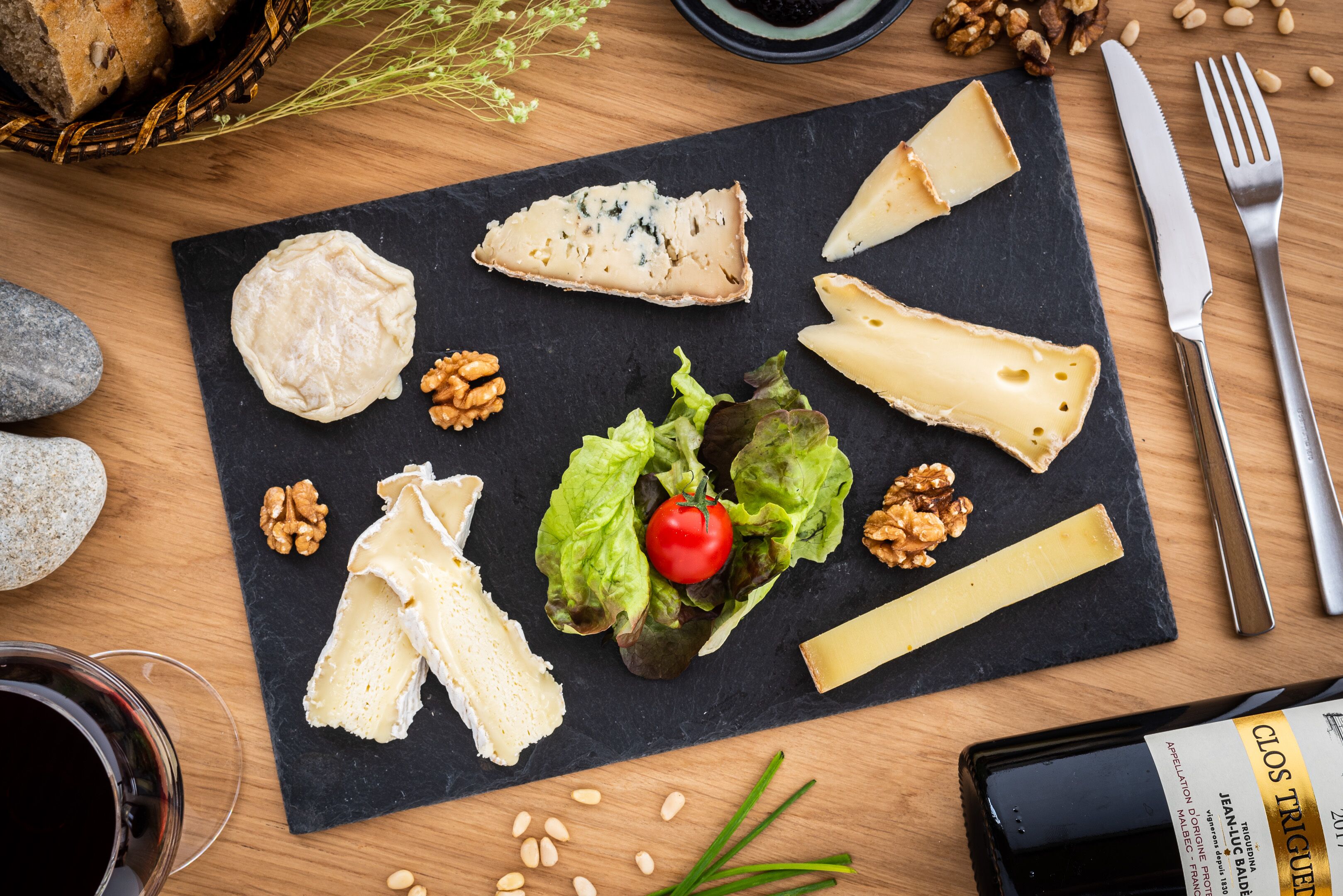 A cheese board with various cheeses, walnuts, and a cherry tomato on a slate board.