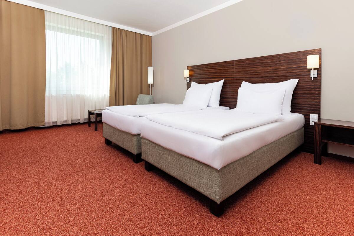Uma cama de casal com cabeceira de madeira, duas mesas de cabeceira, uma cadeira e uma janela grande com cortinas.