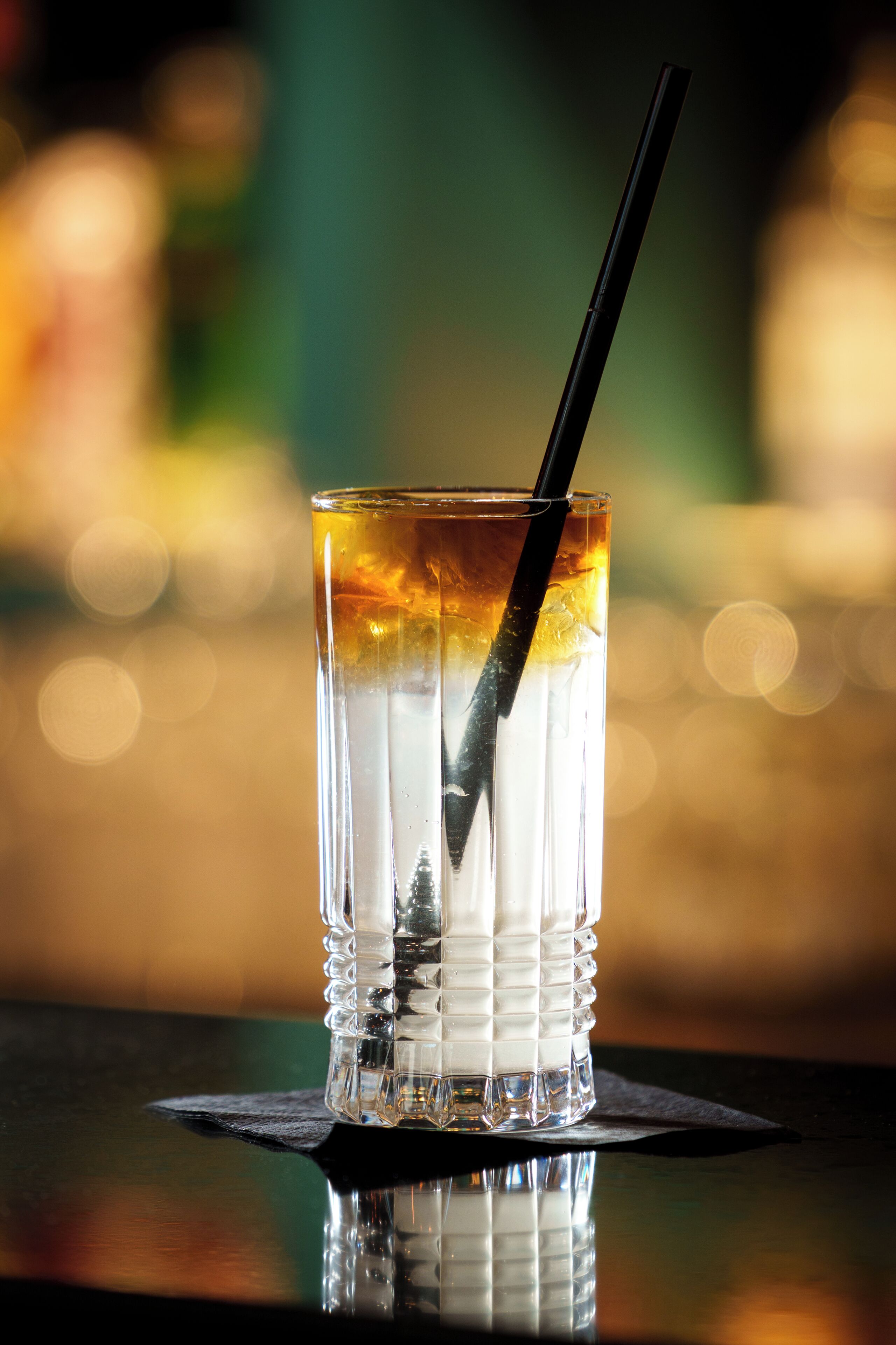A clear glass with a straw, ice cubes, and a golden liquid, possibly a cocktail, on a reflective surface with a blurred background.