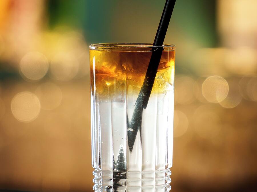 Um copo de vidro transparente com gelo, canudo e líquido dourado, possivelmente um cocktail, sobre uma superfície reflexiva com fundo desfocado.