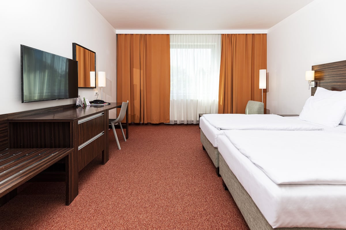 Quarto de hotel com uma cama, escrivaninha, cadeira, televisão e janela com cortinas.