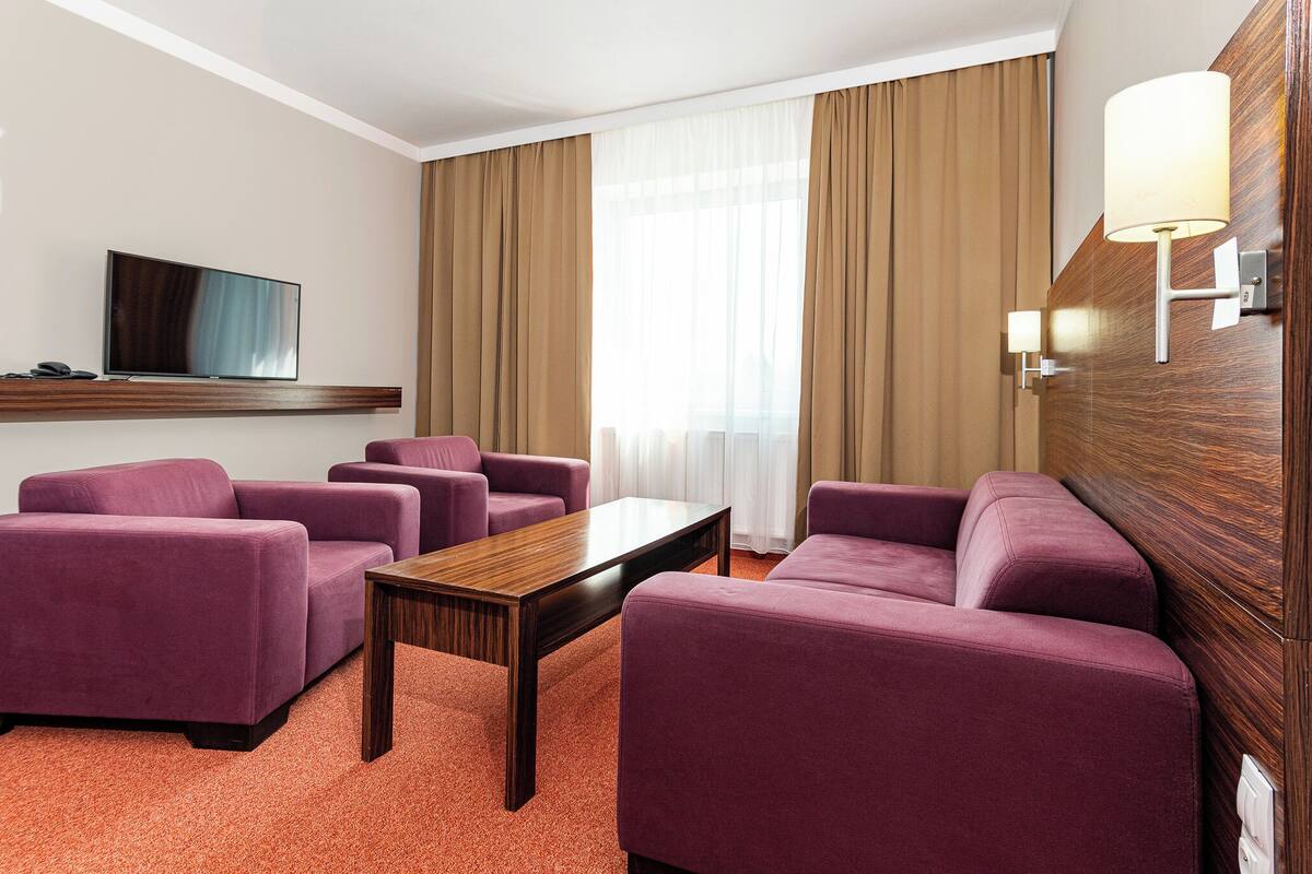 Quarto de hotel com televisão de tela plana, mesa de centro de madeira, sofás roxos, parede de destaque em madeira e uma grande janela com cortinas.