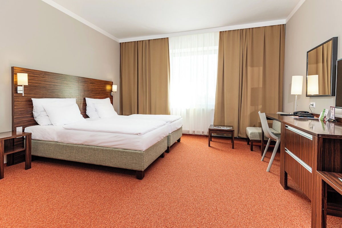 Quarto de hotel com cama, escrivaninha, cadeira e uma janela grande com cortinas.
