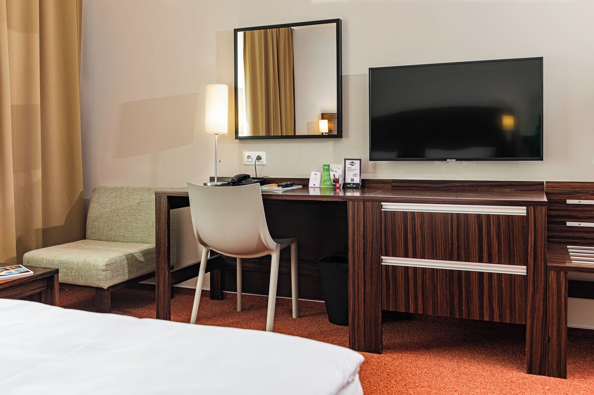 A hotel room with a bed, a desk with a chair, a TV, a lamp, and a trash bin.