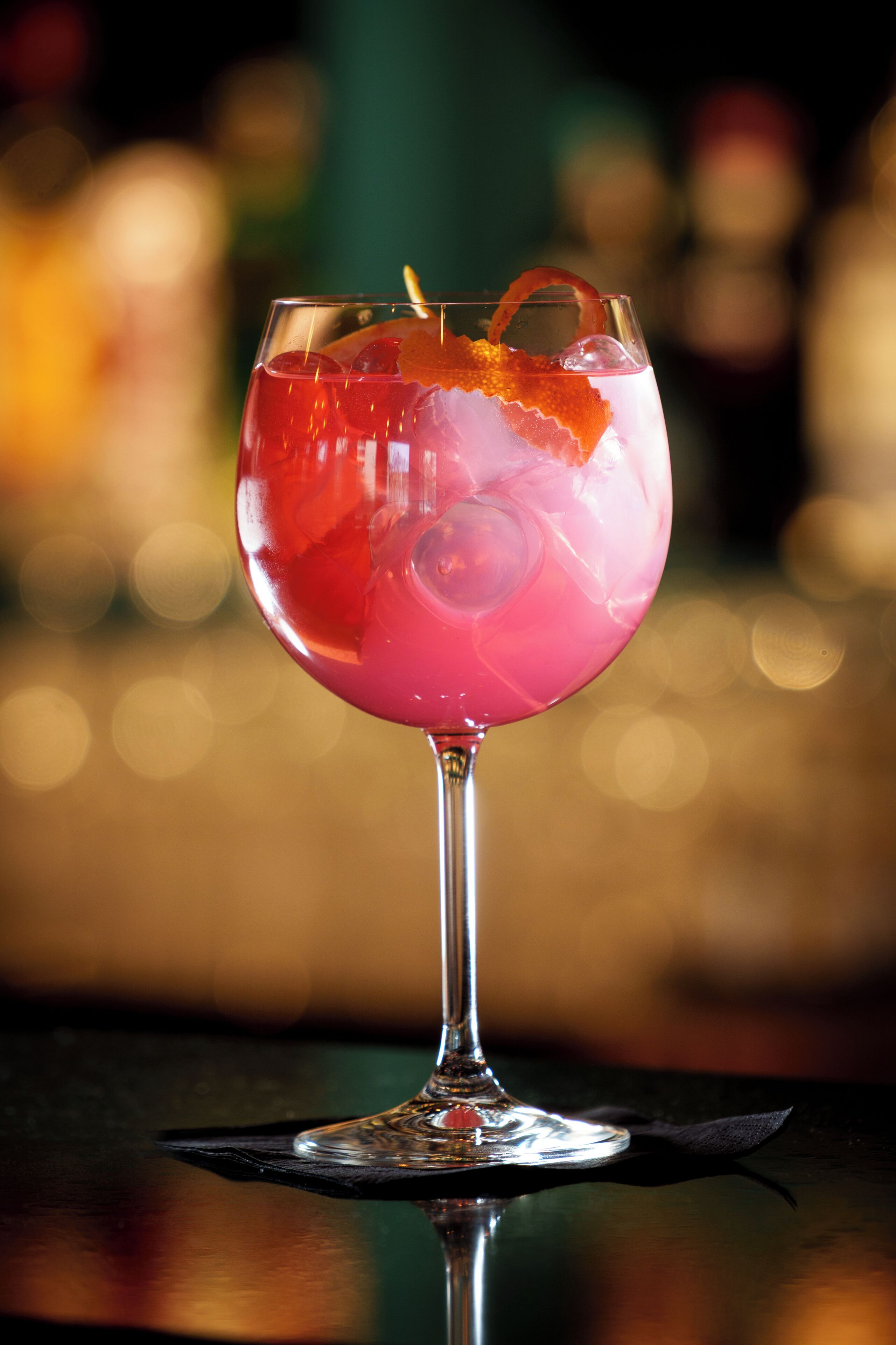 Un verre à cocktail contenant une boisson rose, des glaçons et une tranche d’orange en guise de décoration, le tout posé sur une surface réfléchissante.