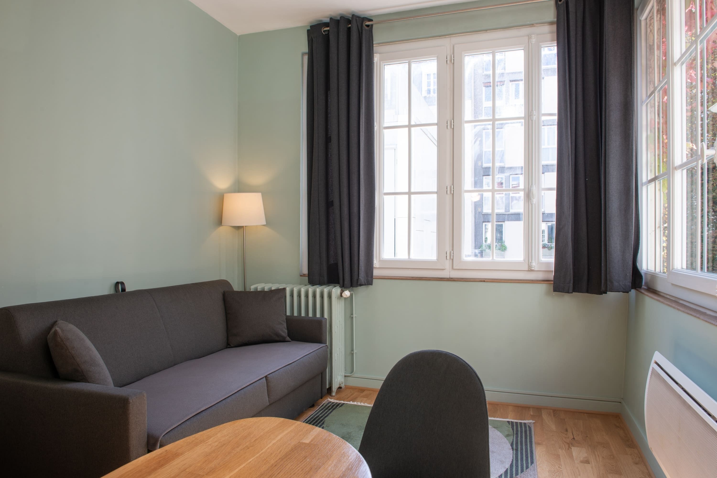 Ein Zimmer mit einer Couch, einem Tisch, einer Lampe, einem Heizkörper und einem Fenster mit Vorhängen.