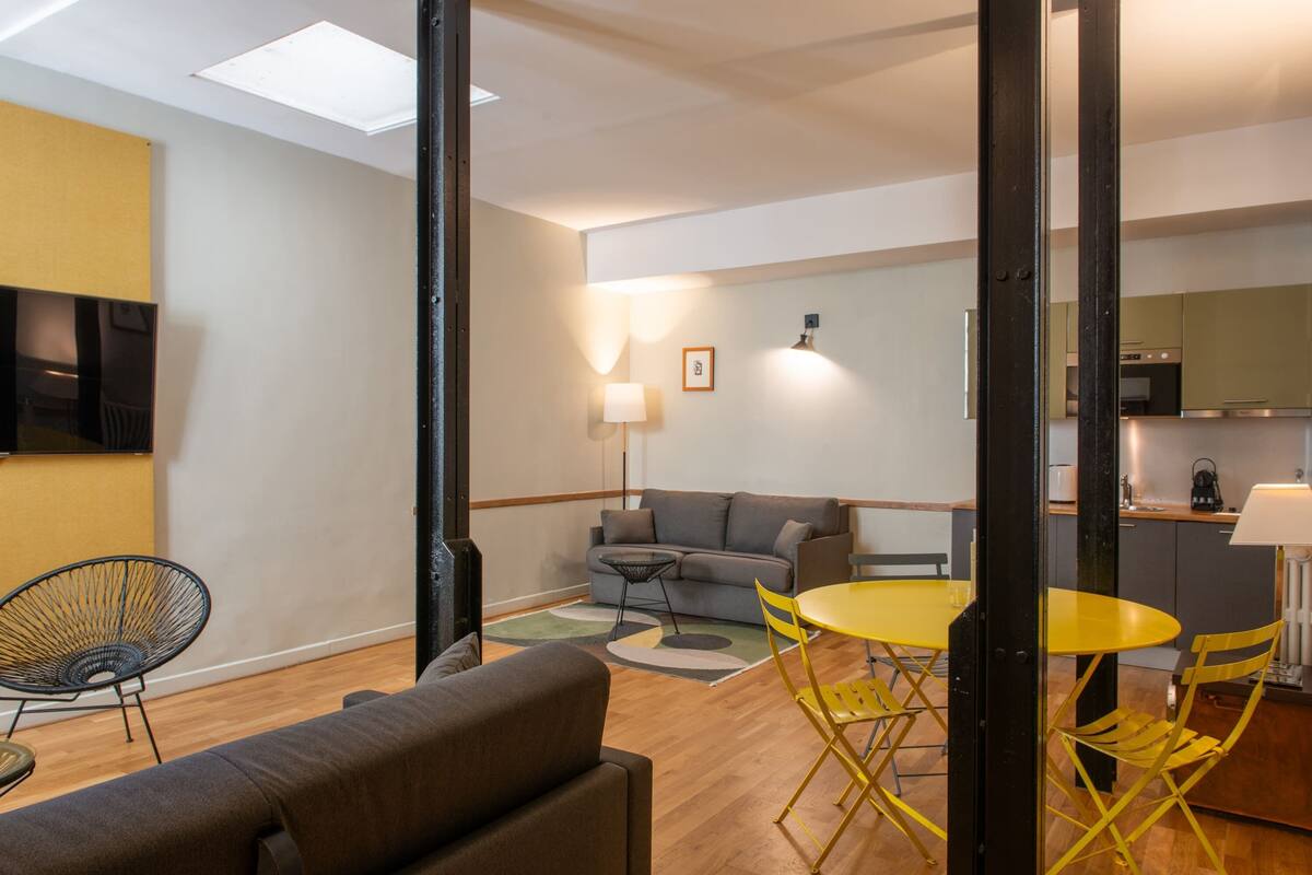 A modern living room with a sofa, yellow chairs, and a small table. There is a kitchen area in the background with cabinets and a stove.