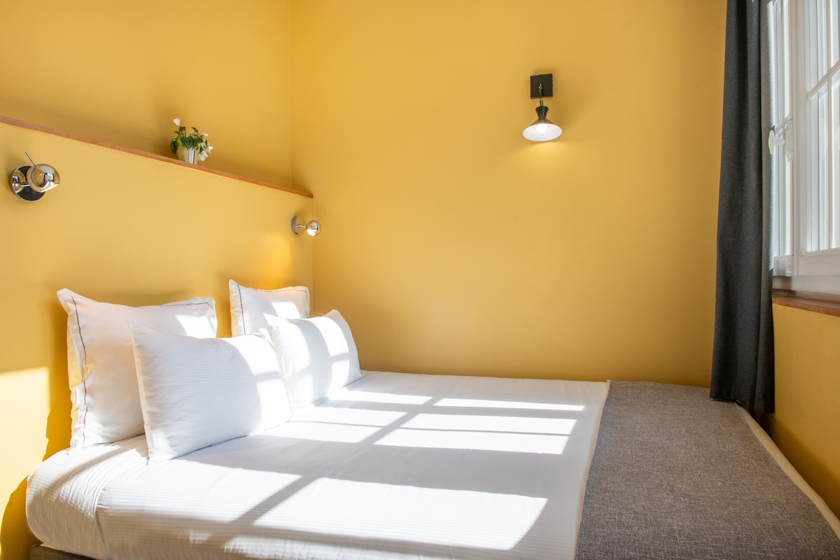 Una cama con ropa blanca, un estante con una planta y una lámpara fijada a la pared.