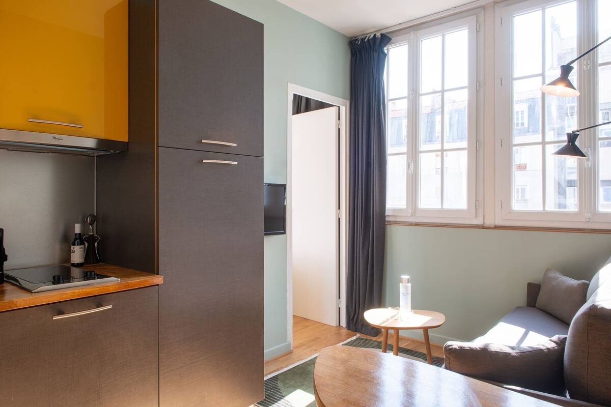 A compact kitchen with a built-in stove, a small table, and a sofa in a room with large windows and a view of buildings.
