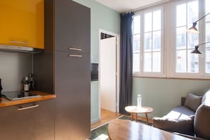 A compact kitchen with a built-in stove, a small table, and a sofa in a room with large windows and a view of buildings. - Hôtel Helzear Montparnasse (Paris)